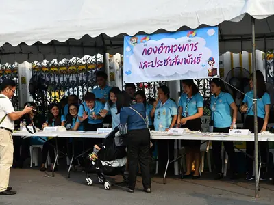 Vrijwilligers in blauwe shirts bij een onthaalbalie onder een banner tijdens het Kinderdag-evenement bij Government House
