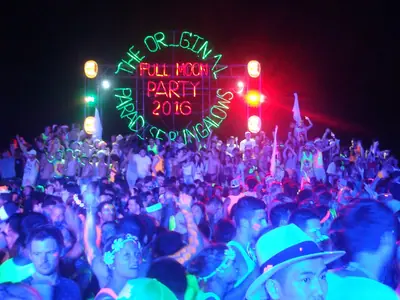 A crowd dancing on the beach at night under a green neon 'Full Moon Party' sign on Koh Phangan (2016)