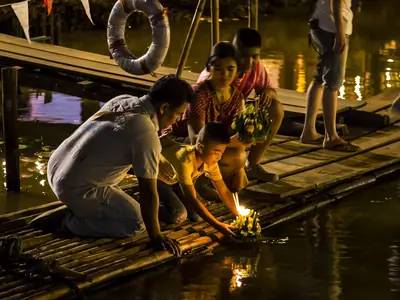 People kneeling on a wooden pier set candle-lit krathongs on the Ping River at night during Loy Krathong (Chiang Mai, same river)