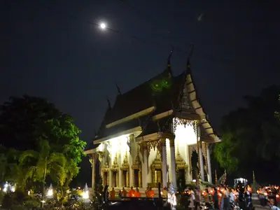 Temple hall lit up at night under a full moon with a crowd gathered during Magha Puja
