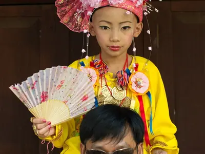 A boy in an elaborate costume and flower headdress carried on a man's shoulders at Poi Sang Long