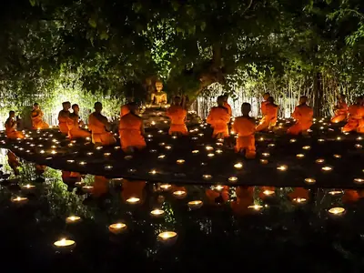 Devotees praying by candlelight on Visakha Bucha night in Thailand, with monks in orange robes in the background