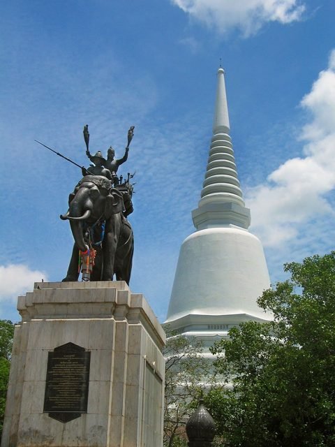 Don Chedi-monument in Suphan Buri: een bronzen beeldengroep op een sokkel voor een witte chedi tegen een blauwe lucht