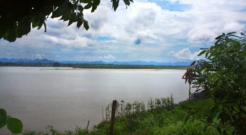De brede Mekong bij Nakhon Phanom onder een gedeeltelijk bewolkte lucht, met bladeren op de voorgrond